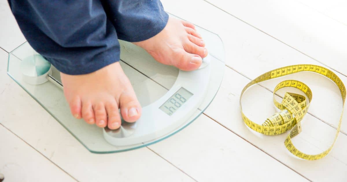 child in blue pants stands on scale with bare feet.